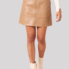 A person wearing a high-waisted camel tan faux leather mini skirt, paired with a white ribbed long-sleeve top and cream ankle boots against a plain background.